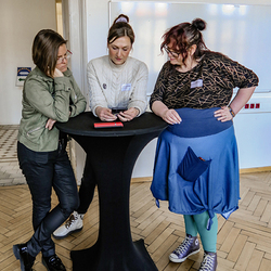 Drei Frauen stehen um einen Stehtisch und schauen gemeinsam auf ein Tablet, während sie eine digitale Schnitzeljagd bearbeiten.