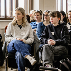 Zwei junge Frauen sitzen aufmerksam im Publikum einer Veranstaltung; im Hintergrund weitere Zuhörende.