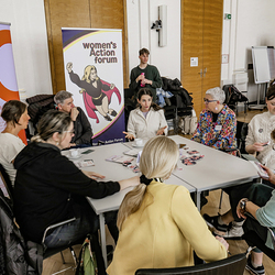 Teilnehmende diskutieren an einem Infotisch des Grazer Frauenrats und Women*s Action Forums über Gleichstellung. Im Hintergrund Banner mit Logos.