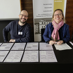 Zwei Personen sitzen an einem Empfangstisch mit vielen Namensschildern. Sie lächeln. Im Hintergrund ein Infoposter zum Studium.