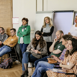 Personen stehen und sitzen aufmerksam in einer Diskussionsrunde – Teilnehmende des Publikums hören zu.