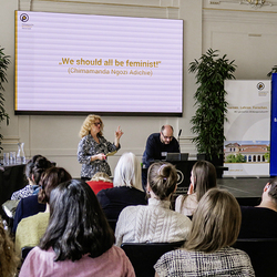 Vortrag mit der eingeblendeten Folie „We should all be feminists“ (Zitat von Chimamanda Ngozi Adichie); Rednerin spricht, Publikum hört zu.