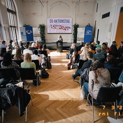 Ein großer, heller Saal mit Publikum, das einer Rednerin an einem Pult zuhört. Auf der Leinwand hinter ihr steht „EU-Aktionstag“ mit abgebildeten EU-Flaggen. Rund 60 Personen sitzen in Reihen, es gibt Infostände und Banner von u. a. UNESCO und dem EU
