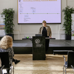 Susanne Kink Hampersberger hält an der PH Steiermark einen Vortrag zu historischen Meilensteinen der Gleichstellung. Auf der Leinwand ist eine Zeitachse mit Stationen wie Aufklärung, Schulpflicht und 20. Jahrhundert zu sehen. Das Publikum folgt aufme
