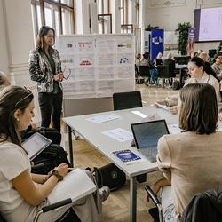 Anneliese Lengger von der Bildungsdirektion Steiermark steht neben einem Tisch mit mehreren Teilnehmerinnen, die aufmerksam zuhören. Sie stellt ihre Arbeit zur reflexiven Pädagogik und Gleichstellung vor. Hinter ihr ist ein Plakatständer mit visualis