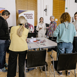 Mehrere Frauen im Gespräch am Infotisch des Women*s Action Forums und des Grazer Frauenrats. Im Hintergrund Roll-ups mit Logos.