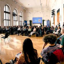 Publikum in einem großen, hellen Raum nimmt an einer Fishbowl-Diskussion teil. Zwei leere Stühle stehen in der Mitte des Kreises, viele Teilnehmende hören aufmerksam zu.