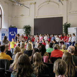 Ein Kinderchor der Praxisvolksschule singt vor Publikum in einem Festsaal, begleitet von zwei Chorleiterinnen.