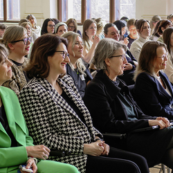 Mehrere Frauen sitzen lächelnd im Publikum und folgen einer Veranstaltung in einem hellen Raum mit großen Fenstern.