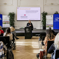 Aleksandra Wierzbicka steht am Rednerpult der PH Steiermark und hält einen Vortrag zur Gleichstellung in der Elementarpädagogik. Auf der Leinwand hinter ihr ist eine Präsentationsfolie mit dem Titel „Elementarpädagogik – Gemeinden fördern Kinderbetre