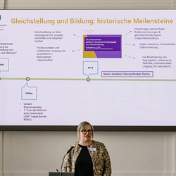 Susanne Kink-Hampersberger steht am Rednerpult der PH Steiermark. Hinter ihr ist eine Präsentationsfolie mit dem Titel „Gleichstellung und Bildung: historische Meilensteine“ zu sehen. Die Folie zeigt eine Zeitachse mit Stationen von 1995 bis 2018, da