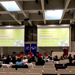 Blick auf das Publikum im Hörsaal,  im  Hintergrund zwei Leinwände auf der das Aktionstage Logo zu sehen ist.